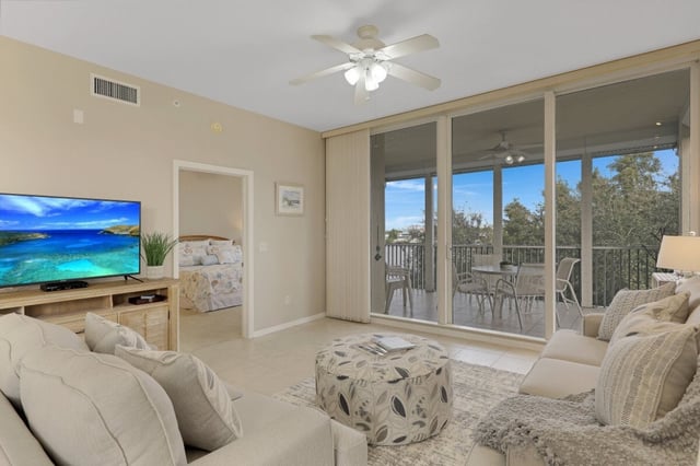Bright condo living room with ceiling fan, flat-screen TV and sliding glass doors opening to a screened balcony with treetop views