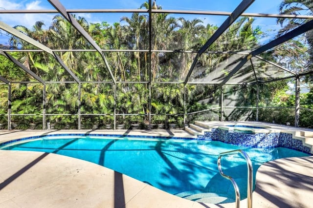 Screened lanai pool with attached spa and blue tile trim, framed by tall palms and tropical greenery