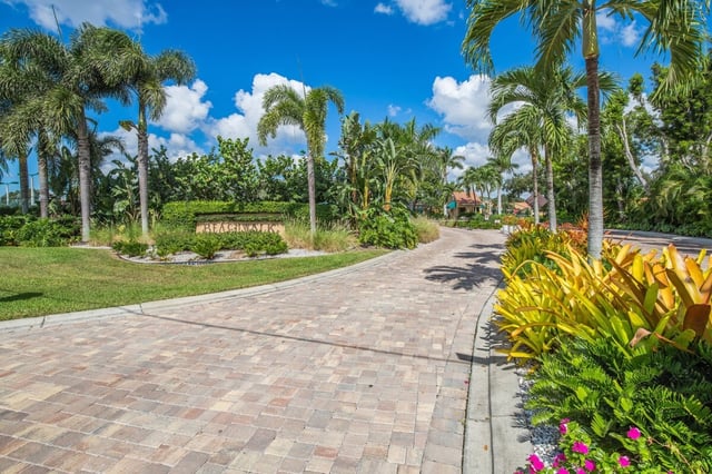 Paver driveway curving past royal palms and tropical plantings toward a Beachwalk entrance sign under blue sky