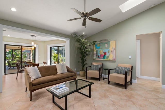 Bright living room with vaulted ceiling, ceiling fan, tan sofa, glass coffee table and tropical plant by the windows