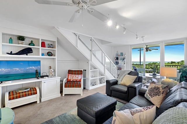 Bright coastal condo living room with white built-in shelves, leather sofa and chairs, staircase, and sliding doors to a water view