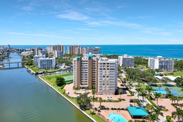 Aerial view of waterfront high-rise condominiums between a calm bay and the blue Gulf, with palms and a pool below