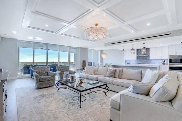 Bright coastal condo living room with coffered ceiling, crystal chandelier, sectional sofa and Gulf views beyond glass doors