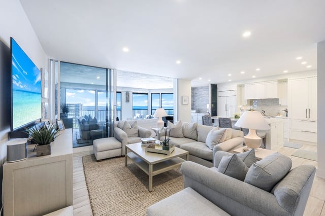 Bright coastal condo living room with gray sofas, jute rug, open white kitchen and Gulf views through sliding glass doors