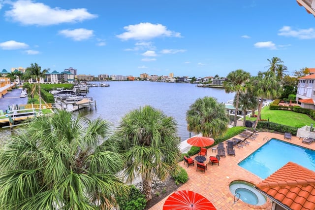 Elevated view of a waterfront pool deck with red umbrellas and palms overlooking a bay lined with docks and boats