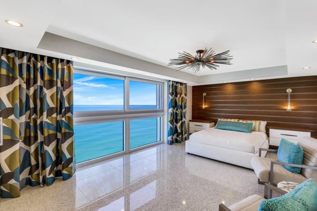 Modern oceanfront bedroom with floor-to-ceiling windows framing a blue Gulf view, wood accent wall and starburst light fixture