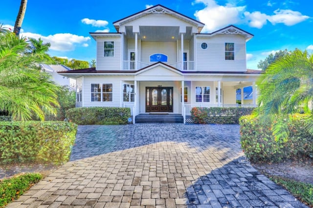 White two-story coastal home with wraparound porch, balcony and paver driveway framed by palms under blue sky