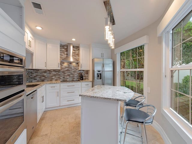 Bright white kitchen with granite island, bar stools, stainless steel appliances and windows overlooking greenery