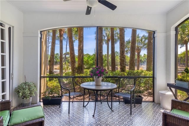 Screened lanai with a bistro table, potted flowers and ceiling fan overlooking palm trees and greenery