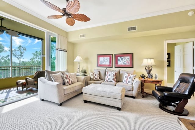 Bright condo living room with cream sofas, palm-leaf ceiling fan and sliding doors to a screened lanai