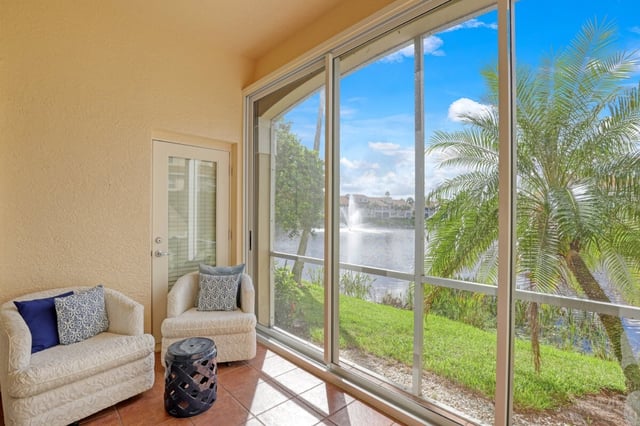 Screened lanai with cushioned armchairs and terracotta tile overlooking a lake with fountain and palm trees