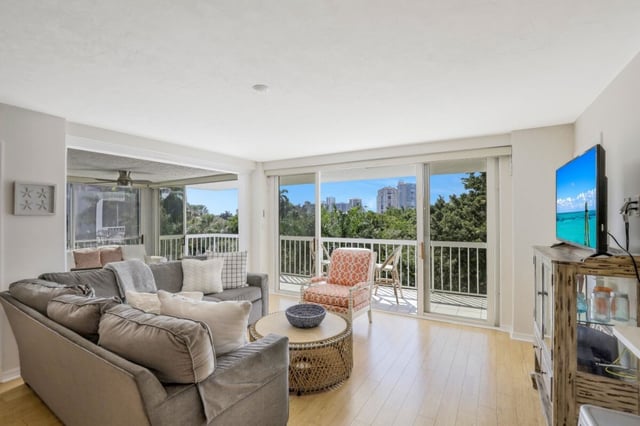 Bright condo living room with gray sectional, wicker coffee table and sliding glass doors opening to a balcony with treetop and skyline views