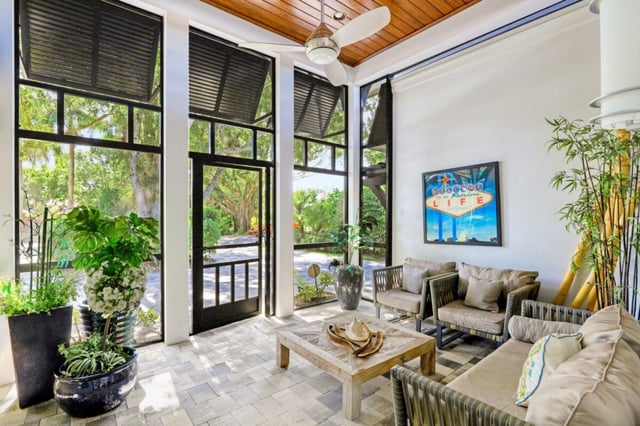 Screened lanai with wood-plank ceiling, ceiling fan, wicker seating, potted plants and framed beach-sign art