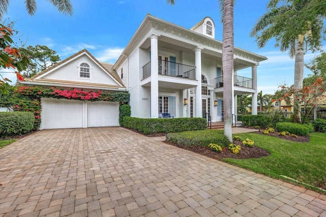 White two-story coastal home with columned balconies, paver driveway, palms and pink bougainvillea under blue sky