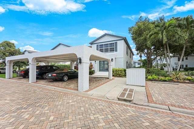 White two-story condo building with covered carport, parked cars, palm trees and brick paver drive under blue sky