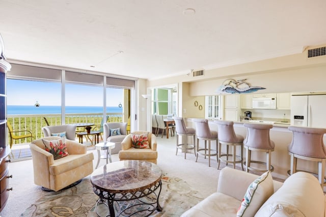 Bright condo living room with floor-to-ceiling windows overlooking the Gulf, armchairs, and a kitchen bar with tall stools