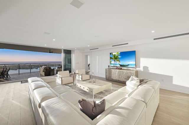 White leather sectional in a bright modern living room with a large TV and balcony overlooking water at sunset