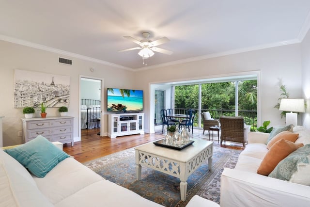 Bright coastal living room with white sofas, teal and orange pillows, ceiling fan, TV, and sliding doors to greenery