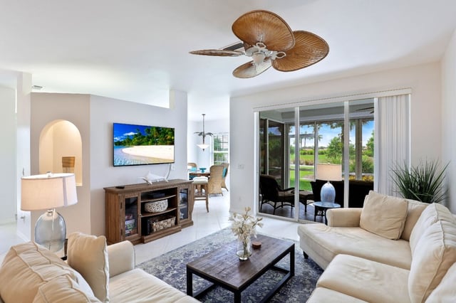 Bright living room with cream sofas, palm-leaf ceiling fan, wall-mounted TV and sliding doors to a screened lanai