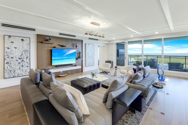Modern condo living room with sectional sofas, wall-mounted TV and floor-to-ceiling windows overlooking greenery and sky
```