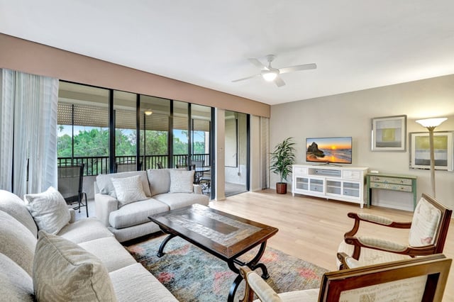 Bright condo living room with sectional sofa, wood coffee table and sliding doors to a screened lanai with green treetop views