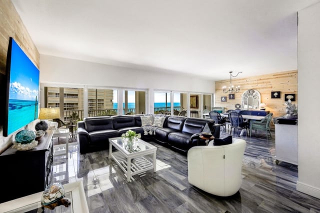 Open-plan condo living room with black leather sofas, white armchair and dining area framing a Gulf view through sliding glass doors