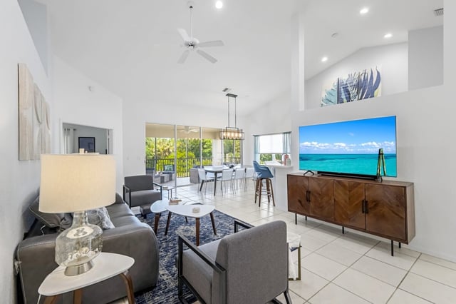 Bright open-concept living room with vaulted ceilings, ceiling fan, large TV on a wood console and sliding doors to a screened lanai