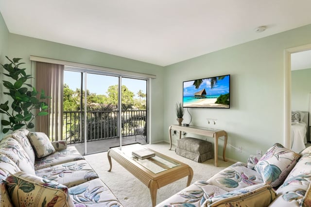 Condo living room with floral sofas, wall-mounted TV, and sliding glass doors opening to a palm-shaded balcony