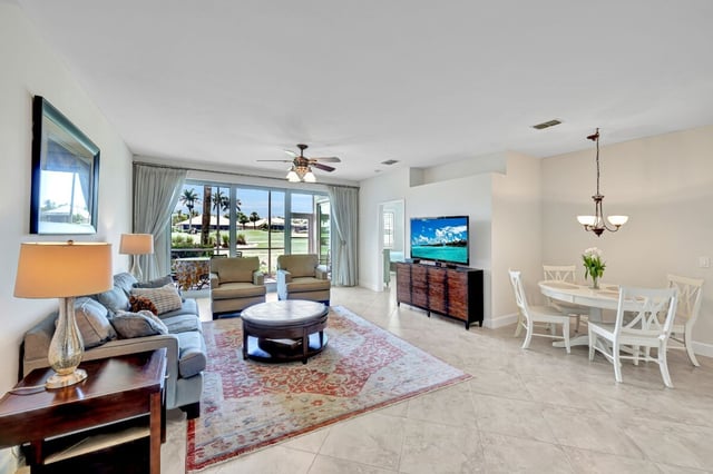 Bright condo living room with tiled floors, ceiling fan, dining set and sliding doors opening to palm-lined greenery