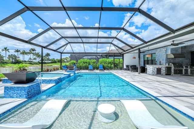 Screened lanai pool with blue mosaic tile, water bowl fountains, sun-shelf loungers and an outdoor kitchen under blue sky