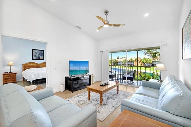 Bright condo living room with pale blue sofas, ceiling fan, and sliding doors opening to a screened lanai and lake view
