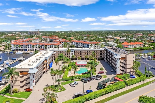 Aerial view of waterfront condo buildings with a pool, palm trees and a marina full of boats under blue sky