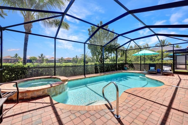 Screened lanai with a kidney-shaped pool, raised spa, brick paver deck and teal umbrella under blue sky