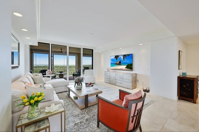 Bright condo living room with white sectional, orange armchairs, wall-mounted TV and balcony overlooking greenery