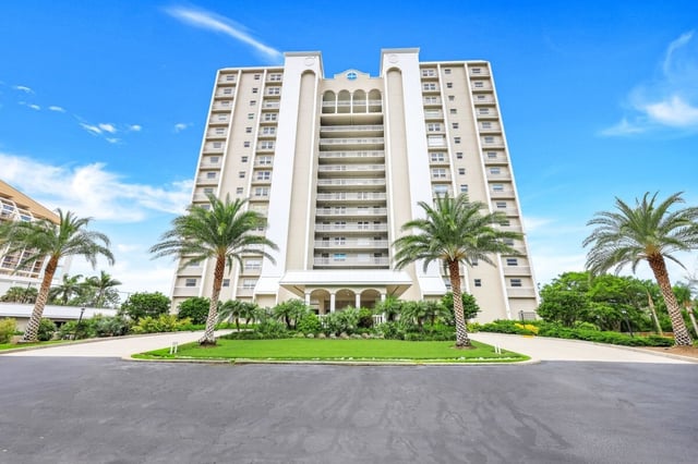 High-rise condo tower with balconies, framed by palm trees and a circular drive under a bright blue sky