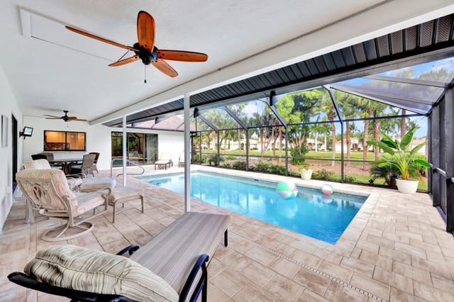 Screened lanai with a blue pool, beach balls, ceiling fans and cushioned loungers overlooking palm-lined greenery
