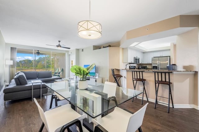 Open-plan condo living area with glass dining table, white chairs, gray sectional sofa and kitchen breakfast bar with stools