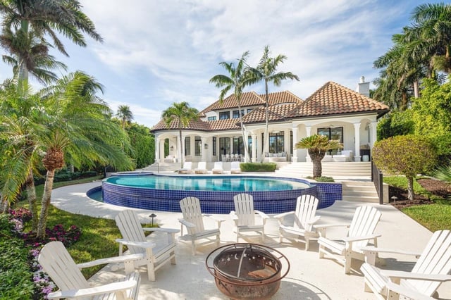 Luxury waterfront estate with blue-tiled infinity pool, white Adirondack chairs around a fire pit and swaying palms