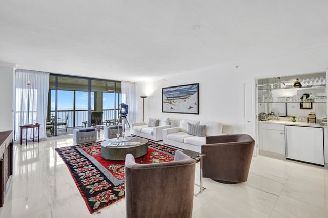 Bright condo living room with white sofas, velvet swivel chairs, patterned rug, wet bar, and Gulf views through sliding doors