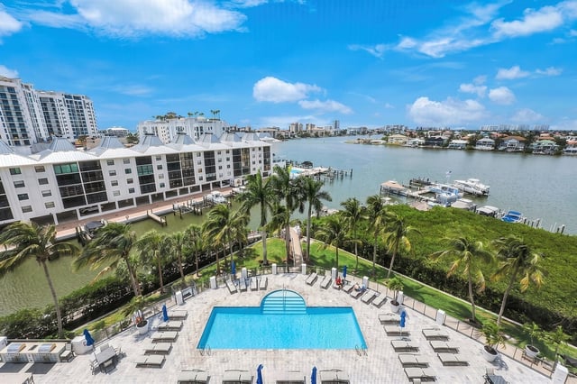 Aerial view of a waterfront condo pool deck lined with palms, loungers and blue umbrellas beside a bay with docked boats