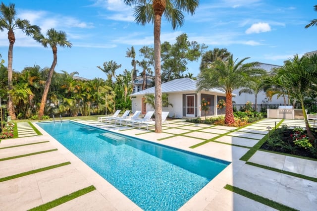Long rectangular pool framed by pale stone pavers, palm trees and a white poolside guest house under blue sky