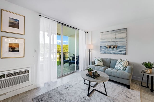 Bright condo living room with gray sofa, sailboat artwork, and sliding doors to a balcony with greenery views