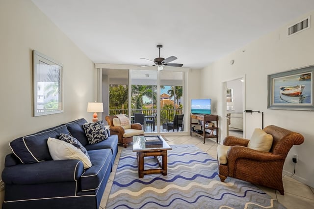 Condo living room with navy sofa, wicker armchairs, ceiling fan and sliding doors to a palm-lined balcony
