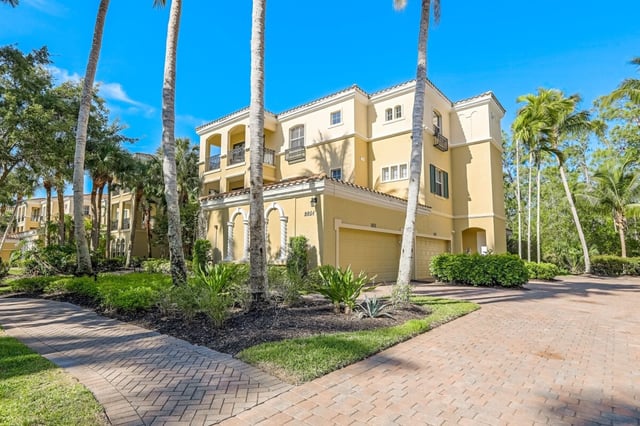 Yellow Mediterranean-style condo building with tile roof, palm trees and a brick paver driveway under blue sky