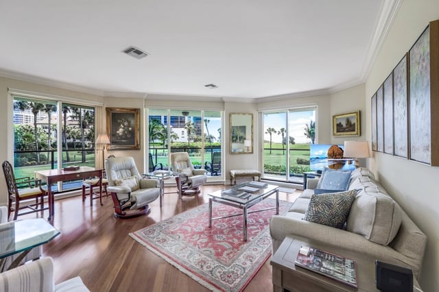 Bright condo living room with hardwood floors, floral area rug, leather recliners and windows framing palm trees