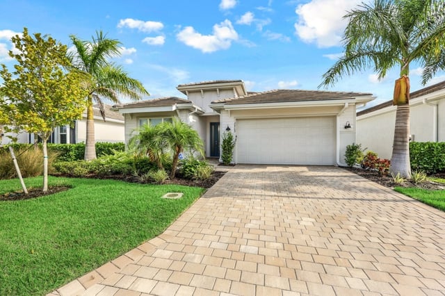Single-story stucco home with a two-car garage, paver driveway, palm trees and green lawn under blue sky
