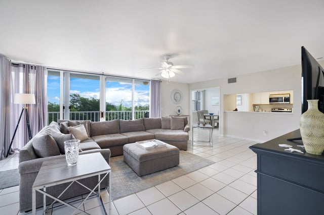 Bright condo living room with gray sectional sofa, ceiling fan, tile floors and balcony windows framing treetop views