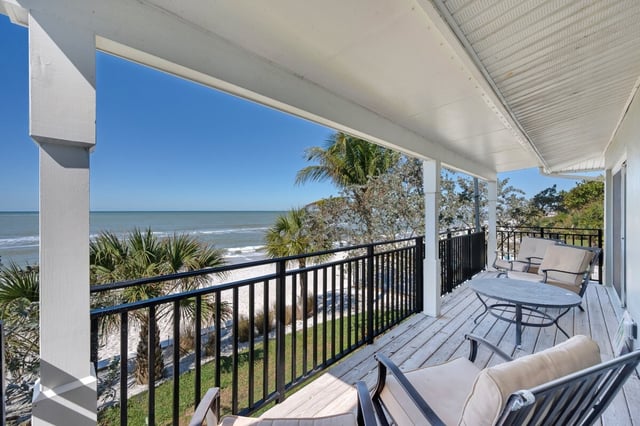 Covered beachfront porch with cushioned patio furniture overlooking palms, white sand and the Gulf under blue sky