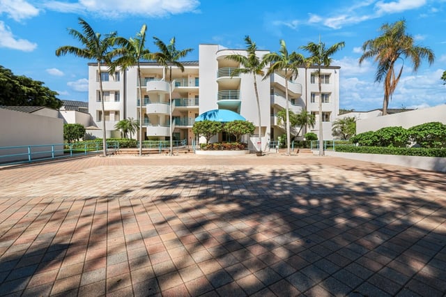White four-story condo building with curved balconies, palm trees and a teal umbrella above a brick paver courtyard