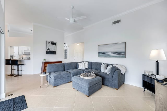 Bright living room with gray sectional sofa, ottoman, tile floors, ceiling fan and coastal wall art